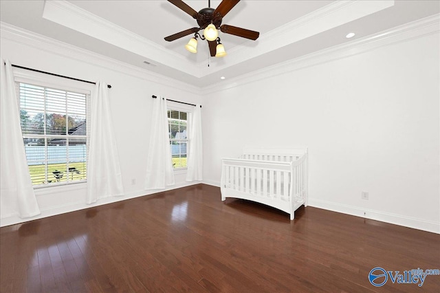 unfurnished bedroom featuring ceiling fan, dark wood-type flooring, crown molding, a tray ceiling, and a nursery area