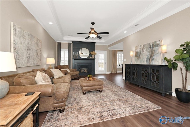 living room featuring a large fireplace, dark hardwood / wood-style floors, a tray ceiling, crown molding, and ceiling fan