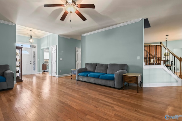 living room featuring stairway, wood finished floors, baseboards, ornamental molding, and ceiling fan