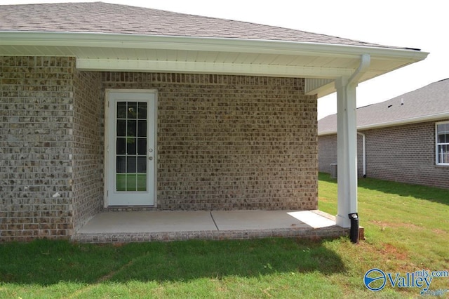 entrance to property featuring a yard, a patio area, brick siding, and roof with shingles