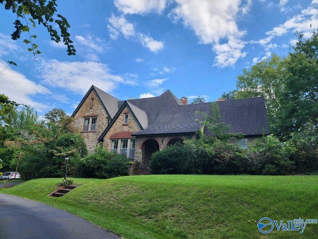 english style home featuring stone siding, a chimney, and a front lawn