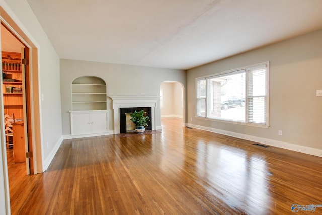 unfurnished living room with visible vents, baseboards, built in shelves, wood finished floors, and arched walkways