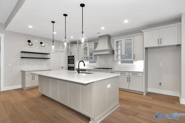 kitchen with an island with sink, custom range hood, light countertops, double oven, and a sink