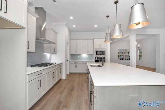 kitchen featuring wall chimney exhaust hood, sink, decorative light fixtures, a kitchen island with sink, and white cabinets