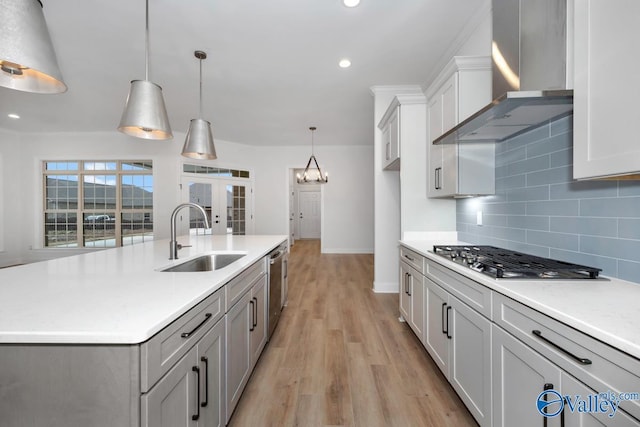 kitchen featuring wall chimney range hood, sink, stainless steel appliances, a center island with sink, and decorative light fixtures