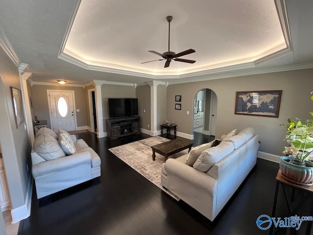 living room featuring crown molding, dark wood-type flooring, ceiling fan, a tray ceiling, and ornate columns