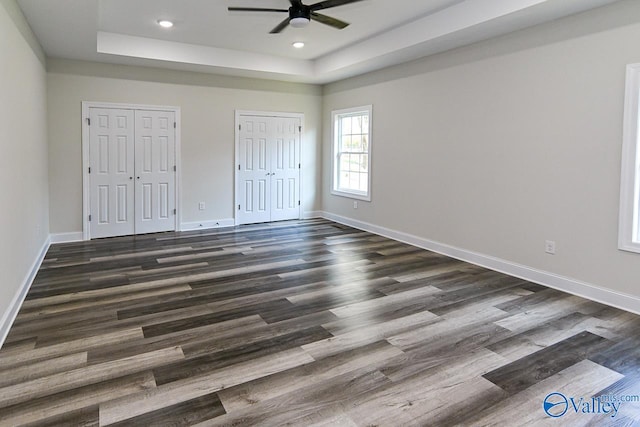 unfurnished bedroom featuring ceiling fan, a tray ceiling, dark hardwood / wood-style floors, and multiple closets