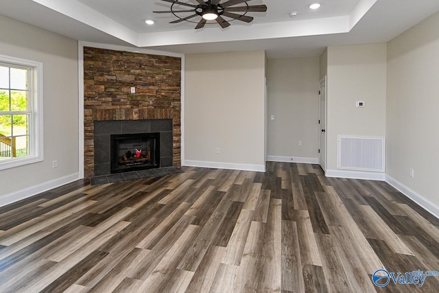 unfurnished living room with a tray ceiling, a stone fireplace, ceiling fan, and dark wood-type flooring