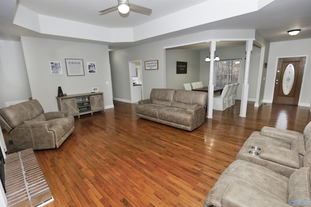 living room with dark wood-type flooring, a tray ceiling, ceiling fan with notable chandelier, and ornate columns