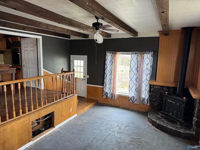 unfurnished living room with wooden walls, a wainscoted wall, ceiling fan, a wood stove, and beam ceiling