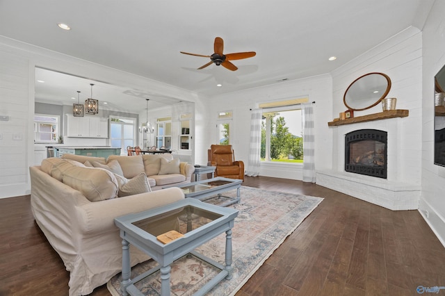 living area with dark wood-style flooring, crown molding, recessed lighting, a fireplace with raised hearth, and ceiling fan with notable chandelier