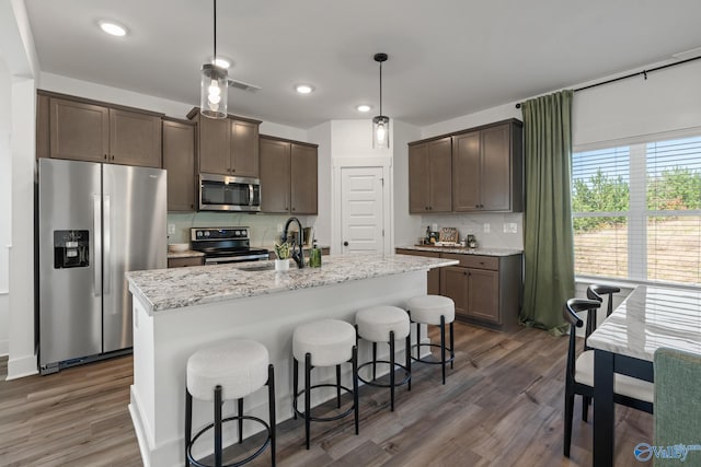 kitchen featuring decorative light fixtures, backsplash, a kitchen island with sink, stainless steel appliances, and light stone countertops