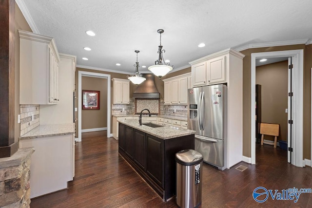 kitchen with pendant lighting, an island with sink, custom exhaust hood, light stone counters, and stainless steel refrigerator with ice dispenser