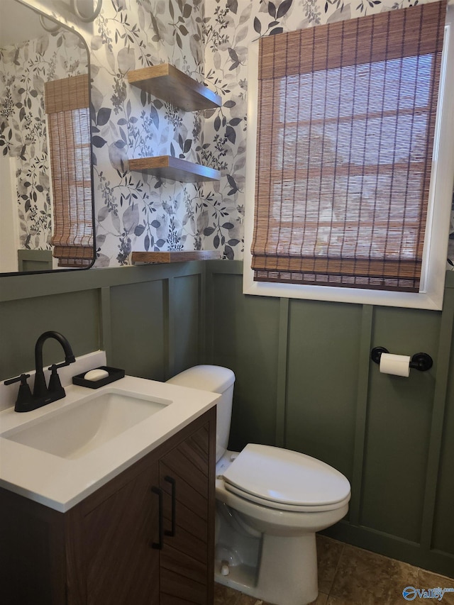 half bathroom with vanity, wallpapered walls, toilet, wainscoting, and a decorative wall
