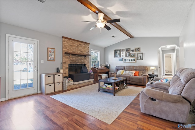 living room with a brick fireplace, a textured ceiling, ceiling fan, wood-type flooring, and lofted ceiling with beams