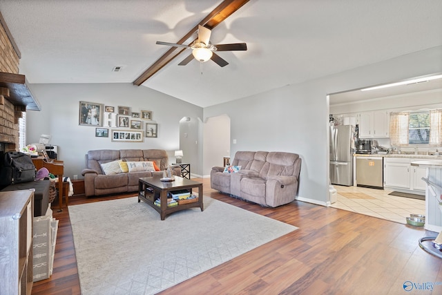 living room featuring vaulted ceiling with beams, light hardwood / wood-style floors, a brick fireplace, and ceiling fan