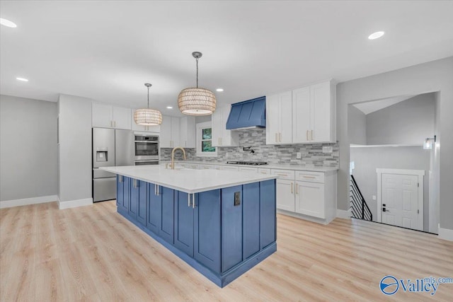 kitchen featuring a center island with sink, custom range hood, appliances with stainless steel finishes, light countertops, and white cabinetry