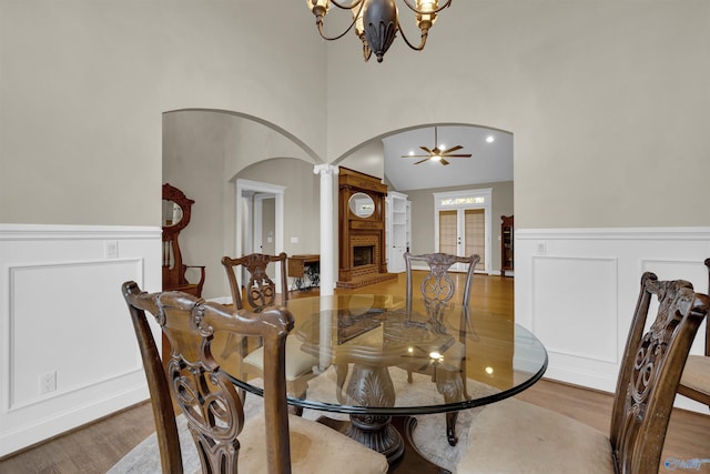 dining space with a towering ceiling, ornate columns, ceiling fan with notable chandelier, wood-type flooring, and a fireplace