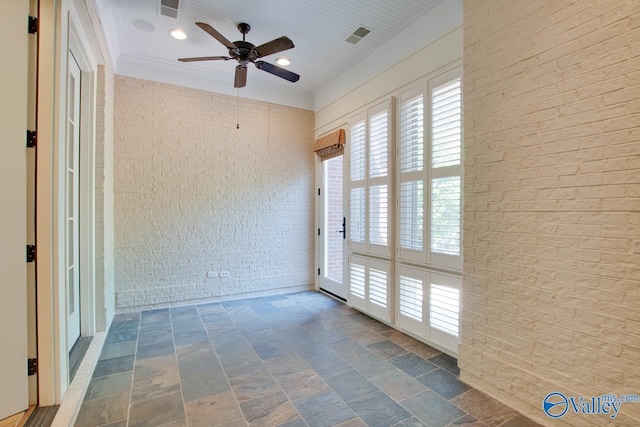 empty room featuring stone finish flooring, brick wall, crown molding, visible vents, and ceiling fan