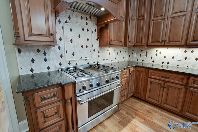 kitchen with light wood finished floors, custom exhaust hood, high end stainless steel range, brown cabinets, and tasteful backsplash