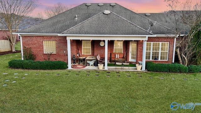 back of house at dusk featuring brick siding, a patio, a shingled roof, and a yard