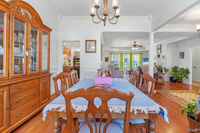 dining area with light wood-style flooring, a wainscoted wall, ceiling fan with notable chandelier, ornate columns, and crown molding