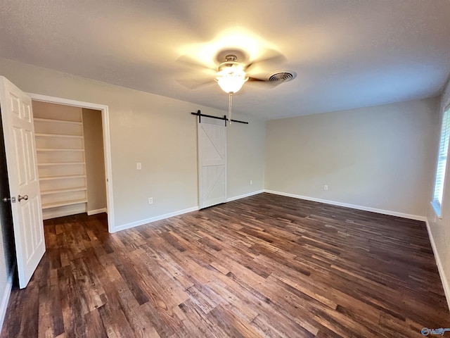 unfurnished bedroom with a ceiling fan, a barn door, dark wood finished floors, baseboards, and visible vents
