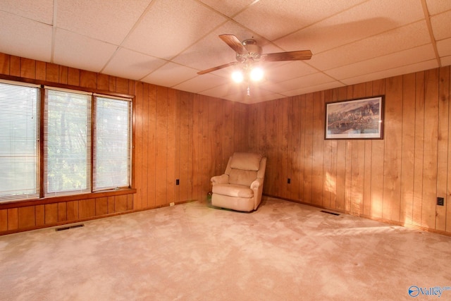 unfurnished room with a drop ceiling, wooden walls, light colored carpet, and ceiling fan