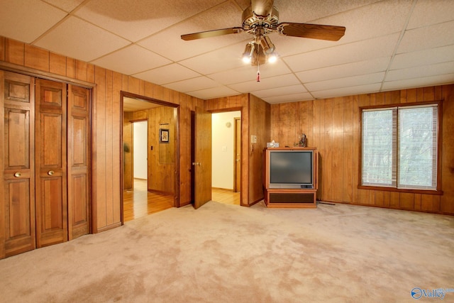 unfurnished living room featuring ceiling fan, a paneled ceiling, wooden walls, and light carpet