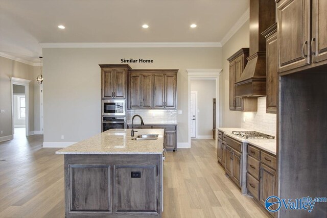 kitchen with wall chimney exhaust hood, sink, appliances with stainless steel finishes, light stone countertops, and a kitchen island with sink