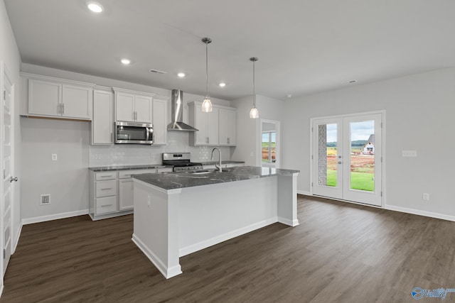 kitchen with wall chimney exhaust hood, a center island with sink, appliances with stainless steel finishes, dark stone counters, and white cabinets