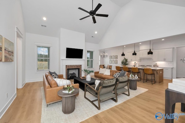 living room featuring a brick fireplace, ceiling fan with notable chandelier, high vaulted ceiling, and light hardwood / wood-style flooring