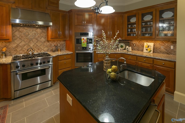 kitchen with appliances with stainless steel finishes, brown cabinetry, a sink, wall chimney range hood, and an island with sink