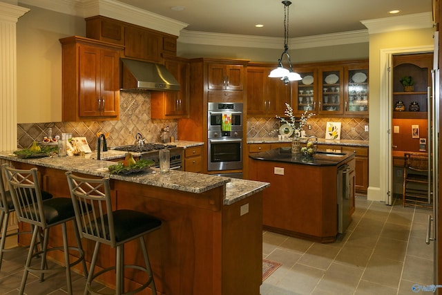 kitchen with a peninsula, stainless steel appliances, a kitchen island, wall chimney exhaust hood, and brown cabinetry