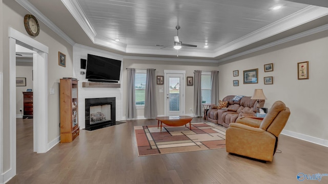 living room featuring wood finished floors, ornamental molding, a ceiling fan, a fireplace with flush hearth, and a tray ceiling