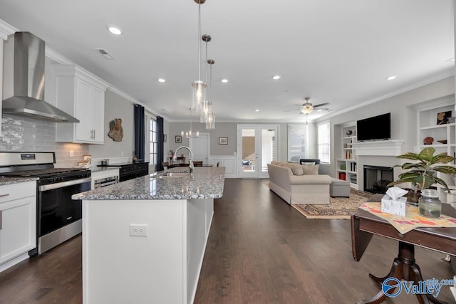 kitchen with sink, wall chimney range hood, stainless steel range oven, dark stone counters, and a center island with sink