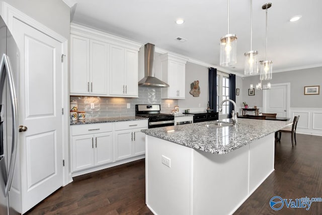 kitchen with appliances with stainless steel finishes, wall chimney exhaust hood, a center island with sink, white cabinets, and hanging light fixtures