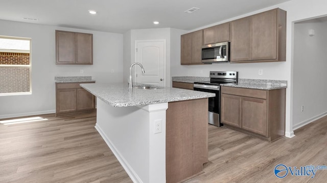 kitchen with light stone countertops, appliances with stainless steel finishes, sink, light hardwood / wood-style flooring, and an island with sink