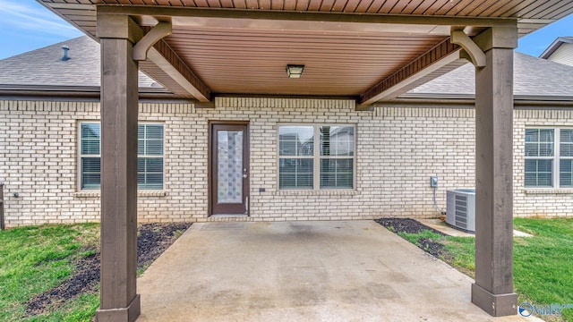 entrance to property with roof with shingles, cooling unit, a patio area, and brick siding