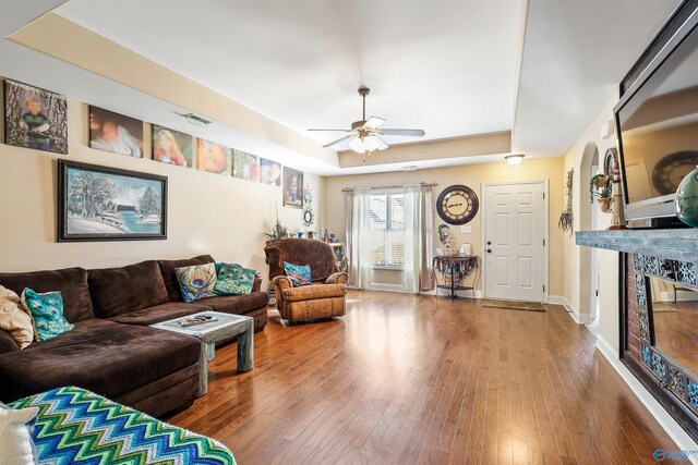 living area with visible vents, a ceiling fan, a tray ceiling, wood-type flooring, and a glass covered fireplace