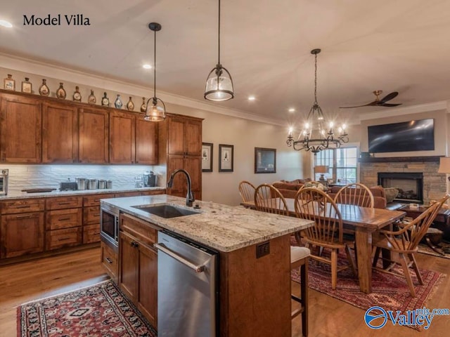 kitchen with stainless steel appliances, light wood-type flooring, ornamental molding, a stone fireplace, and a kitchen island with sink