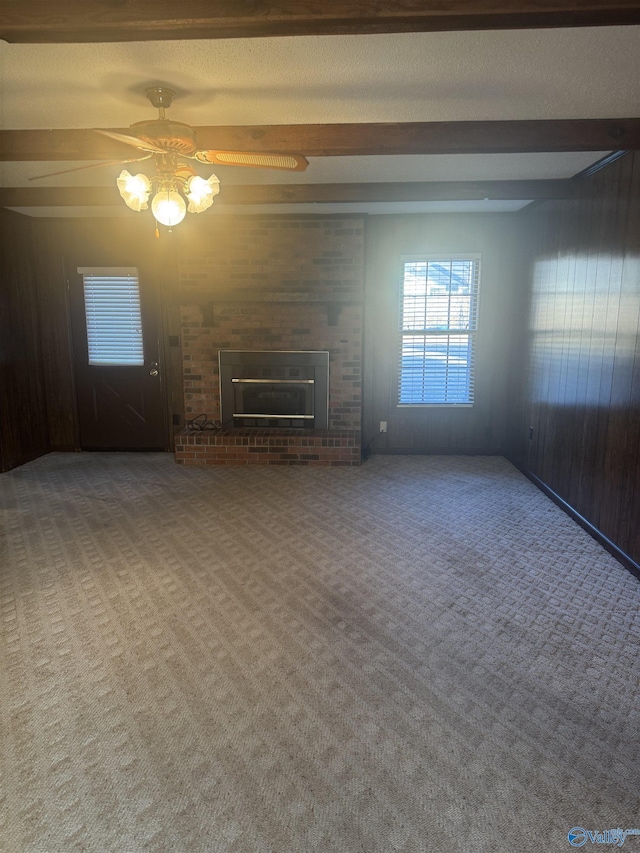 unfurnished living room featuring wooden walls, a brick fireplace, ceiling fan, beamed ceiling, and carpet floors