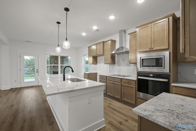 kitchen featuring an island with sink, sink, light stone counters, stainless steel appliances, and wall chimney range hood