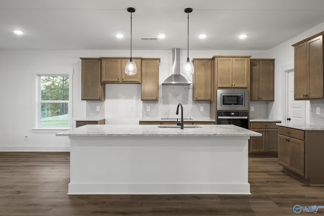 kitchen with wall chimney exhaust hood, sink, stainless steel microwave, an island with sink, and light stone countertops