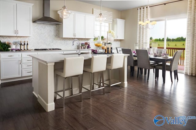 kitchen featuring sink, wall chimney range hood, an island with sink, white cabinets, and decorative light fixtures