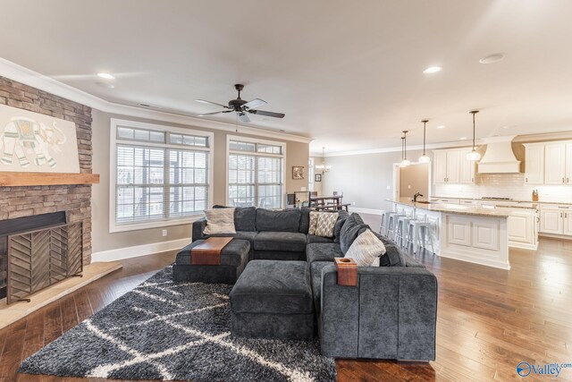 living room with crown molding, ceiling fan with notable chandelier, a large fireplace, baseboards, and dark wood-style floors