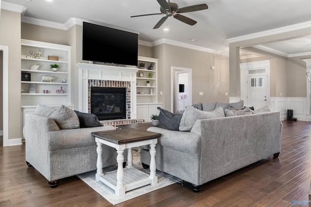 living area with a brick fireplace, crown molding, built in features, wainscoting, and dark wood-style flooring