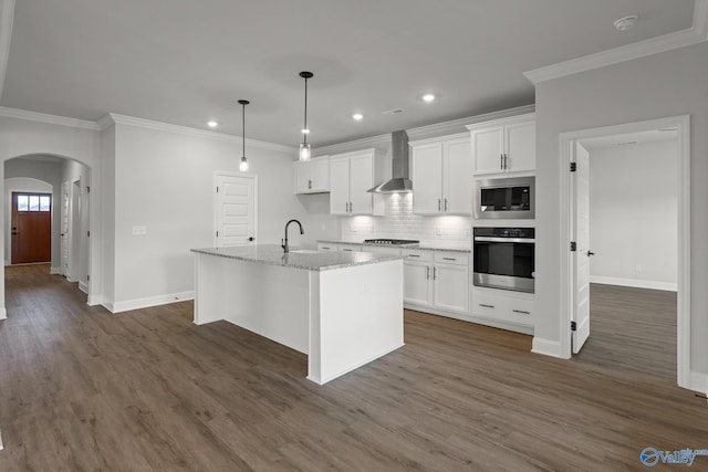 kitchen with dark wood-type flooring, white cabinets, wall chimney exhaust hood, an island with sink, and stainless steel appliances