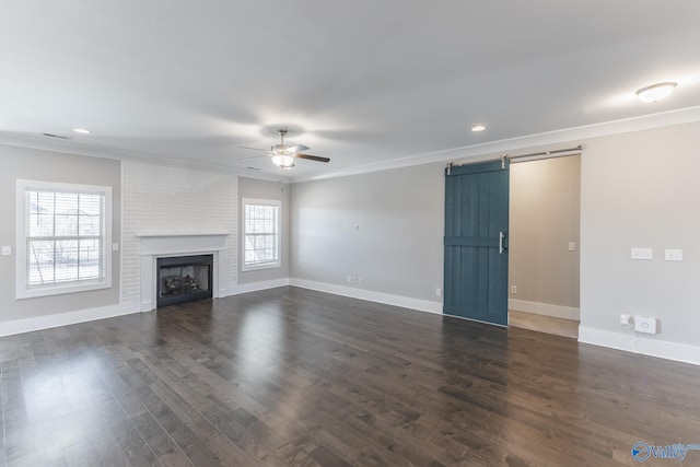 unfurnished living room with ornamental molding, ceiling fan, a barn door, a fireplace, and dark hardwood / wood-style floors