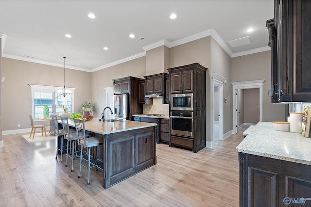 kitchen featuring light wood-type flooring, appliances with stainless steel finishes, light stone countertops, a chandelier, and an island with sink